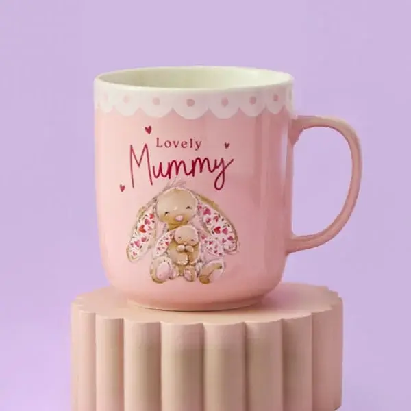 A pink mug with a scalloped white rim featuring an illustration of two cuddly bunnies and the words Lovely Mummy, displayed on a pastel pedestal against a lilac background.